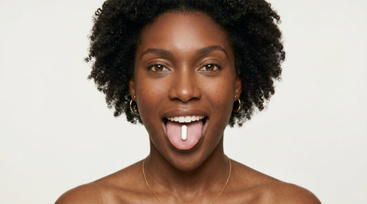 A smiling woman with natural curly hair balancing a single white β-NMN 500mg capsule on her tongue, against a soft neutral background